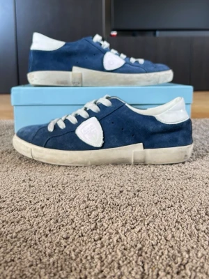 Blå mockasneakers med vita detaljer - Snygga sneakers i blå mocka med vita detaljer och vit sula. Skorna har vita skosnören och en vit hälkappa samt en vit logotyp på sidan. Låg modell som passar perfekt till jeans eller shorts. Klassisk och stilren design.