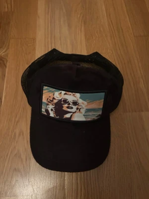 Svart truckerkeps med retrotryck - Snygg svart truckerkeps från Revenite med mesh på sidorna och bak. Framtill finns ett färgstarkt retrotryck av en kvinna med solglasögon mot en strandbakgrund. Justerbar snapback för perfekt passform. Perfekt accessoar för en cool och avslappnad stil.