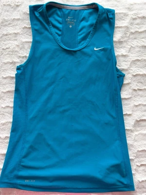 Blått träningslinne från Nike - Snyggt blått träningslinne från Nike med Dri-FIT-material som andas. Linnet har en sportig passform, rund halsringning och diskret Nike-logga på bröstet. Perfekt för gymmet, löpning eller andra sportaktiviteter.