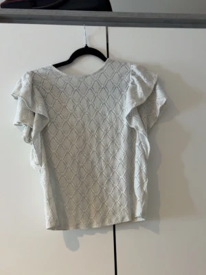 Vit blus med volangärmar - Säljer en söt vit blus med diskret mönster och volangdetaljer på ärmarna. Blusen har rund halsringning och knapp i nacken. Perfekt för dig som gillar en feminin och stilren look.