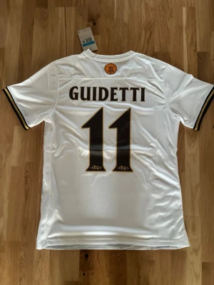 AIK vit matchtröja Guidetti 11 Nike - Säljer en vit AIK matchtröja från Nike med Guidetti och nummer 11 på ryggen. Tröjan har gulddetaljer, AIK:s klubbmärke på bröstet och ett snyggt tryck av Stockholm på framsidan. Korta ärmar med svarta och guldiga ränder vid ärmslut.