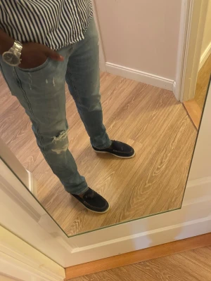 Ljusblå slitna jeans herr - Säljer ett par ljusblå jeans med slitningar och hål på ena knät från Replay. Jeansen har en smal passform och klassisk femficksdesign. Perfekta för en avslappnad och trendig stil. 