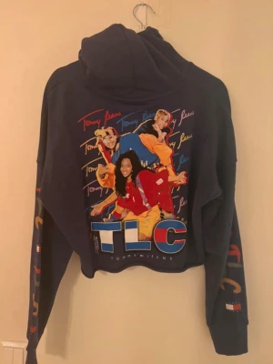 Tommy Jeans TLC hoodie med tryck - Mörkblå hoodie från Tommy Jeans med stort TLC-tryck på ryggen och färgglada detaljer. Framsidan har logga och TLC-tryck. Hoodien har huva och tryck på ärmarna. Perfekt för dig som gillar streetwear och 90-talsvibbar. Endast använd hemma!