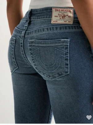 Blå bootcut jeans från True Religion - Säljer ett par klassiska blå bootcut jeans från True Religion med ikoniska kontrastsömmar och stora bakfickor med U-formad brodyr. Jeansen har normal midja och är tillverkade i mjuk denim med lätt stretch för skön passform.
