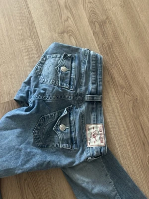 Blå True Religion Joey jeans - Säljer ett par blå True Religion Joey jeans med låg midja och utsvängda ben. Jeansen har dekorativa ficklock med stora silverknappar och tydliga sömmar. Klassisk True Religion-lapp bak och cool tvättad denim-look.