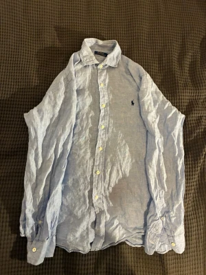 Ljusblå linneskjorta från Polo Ralph Lauren - Säljer en ljusblå skjorta i linne från Polo Ralph Lauren med klassisk krage och knappar framtill. Skjortan har lång ärm och den ikoniska loggan broderad på bröstet. Perfekt för en avslappnad men ändå stilren look.