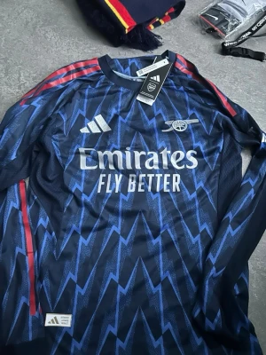 Arsenal långärmad bortatröja Adidas - Säljer en långärmad Arsenal bortatröja från Adidas i mörkblått med blått blixtmönster och röda detaljer på axlarna. Tröjan har tryck med 'Emirates Fly Better' och spelarnamn Gyökeres 14 på ryggen. Perfekt för fotbollsfans!