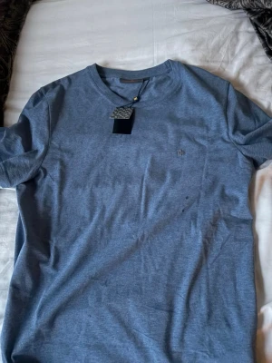 Blå t-shirt från Morris - Snygg blå t-shirt från Morris med diskret logga broderad på bröstet. Klassisk rund halsringning och korta ärmar. Tillverkad i mjuk bomull som känns skön mot huden. Perfekt till jeans eller shorts för en clean look.