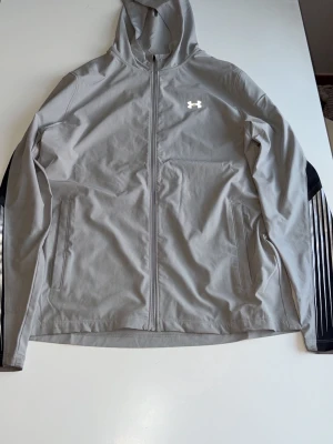Grå vindjacka med huva från Under Armour - Snygg grå vindjacka från Under Armour med hel dragkedja framtill och huva. Jackan har svarta detaljer längs ärmarna och två sidofickor med dragkedja. Perfekt för dig som vill ha en sportig och stilren look.