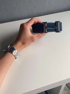 Blått skärp med silverfärgat H-spänne - Snyggt blått bälte med ett stort H-format spänne i silverfärg. Bältet har vita sömmar längs kanterna och är gjort i skinn. Perfekt accessoar för att lyfta din outfit.