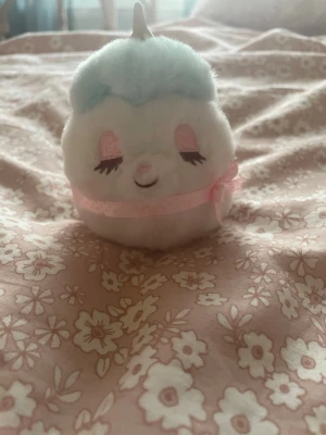 Söt enhörning plushie med pastellfärger - En mjuk och fluffig enhörning plushie i vitt med ljusblå man och svans, rosa detaljer och ett litet horn. Den har broderade ögonfransar och ett rosa band runt kroppen. Perfekt som dekoration eller gosedjur.