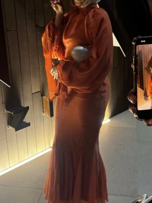 Lång orange klänning med vida ärmar - ENDAST ANVÄNT 1 GÅNG; Säljer en elegant långklänning i en varm orange färg. Klänningen har höghalsad design, vida långa ärmar med knytband vid handlederna och en mjuk, flytande passform. Perfekt för dig som vill sticka ut med en unik och trendig look.