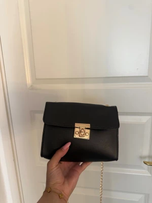 Svart axelväska med guldkedja - Stilren svart axelväska i slätt material med guldfärgad kedja och låsdetalj framtill. Väskan har en fyrkantig form och minimalistisk design som passar till många olika outfits.