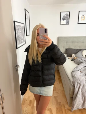 Svart pufferjacka med huva - Säljer en svart pufferjacka med huva och dragkedja framtill. Jackan har breda, vadderade kanaler och en boxig passform som ger en riktigt puffig och trendig look. Perfekt för kalla vinterdagar och enkel att styla till olika outfits.