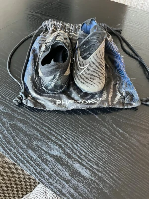 Nike Phantom svart/grå fotbollsskor - Säljer ett par Nike Phantom fotbollsskor i svart och grått med snyggt mönster och vita detaljer. Skorna har blå dobbar och är designade för bra grepp på planen. Levereras med tillhörande svart/blå påse. Perfekta för dig som vill ta spelet till nästa nivå.