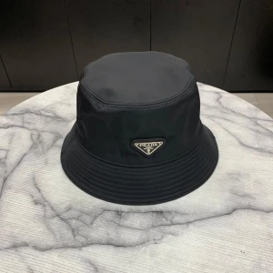 Svart bucket hat i nylon - Snygg svart bucket hati glansigt nylonmaterial. Klassisk form med bred, nedåtlutande brätte och ikoniska metalltriangeln med logga framtill. Perfekt accessoar för en trendig och stilren look.
