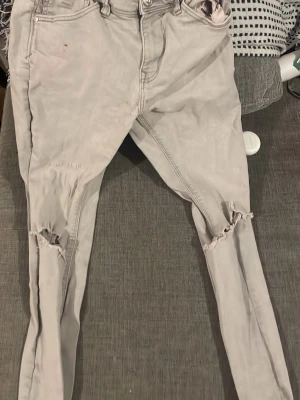 Ljusgrå skinny jeans med slitningar - Säljer ett par ljusgrå skinny jeans med slitna knän och klassisk femficksdesign. Jeansen har låg midja och smal passform som sitter tight hela vägen ner. Perfekta för dig som gillar en trendig och avslappnad stil.