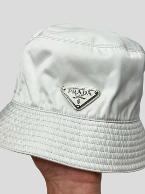 Vit bucket hat från Prada - Snygg vit bucket hat från Prada med den ikoniska metalltriangeln framtill. Hatten är tillverkad i ett glansigt material som ger en lyxig känsla och har klassisk rund form med bred, nedåtlutande brätte. Perfekt accessoar för en trendig streetwear-look.