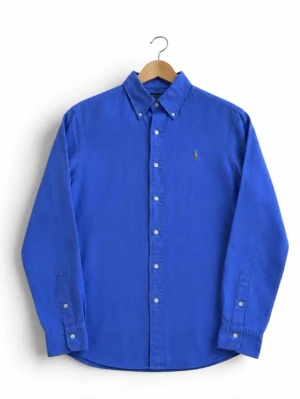 Lila skjorta från Polo Ralph Lauren - Klassisk lila skjorta från Polo Ralph Lauren med button-down krage och broderad logga på bröstet. Skjortan har långa ärmar och vita knappar framtill. Perfekt till jeans eller chinos för en clean och stilren look.