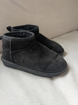 Svarta mockatofflor med foder - Mysiga svarta tofflor i mockaimitation med mjukt foder på insidan. De har en enkel design med rund tå och låg ankel, samt en liten ögla bak för att lätt ta på sig. Perfekta för att hålla fötterna varma hemma under kyliga dagar.