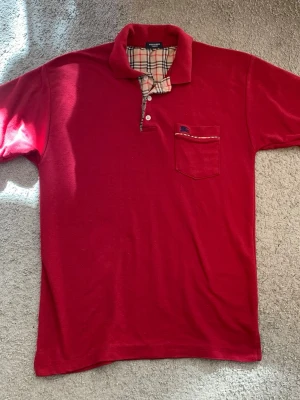 Röd Burberry piké med rutig detalj - sitter som M-L. Snygg röd t-shirt från Burberry med klassisk rutig detalj på insidan av kragen och knappslån. Bröstficka med liten broderad logga och rutigt band. Perfekt för dig som gillar stilrena plagg med en twist.
