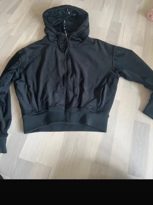Svart hoodie med dragkedja STRONGER - Snygg svart hoodie med dragkedja och huva från STRONGER. Tillverkad i mjukt material med breda muddar och broderad logga på ryggen. Perfekt för en sportig och avslappnad stil.