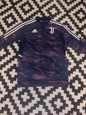 Juventus träningströja från Adidas - Mörkblå Juventus träningströja från Adidas med rosa mönster, vita ränder på axlarna och halv dragkedja. Tröjan har lång ärm och klubbens logga på bröstet. Perfekt för fotbollsträning eller chill. Tillverkad i funktionsmaterial.