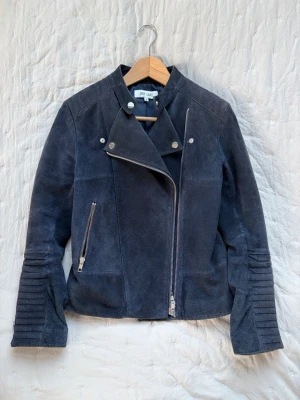 Mörkblå skinnjacka från Dry Lake - Snygg mörkblå skinnjacka från Dry Lake med asymmetrisk dragkedja, dekorativa sömmar på ärmarna och silverfärgade detaljer. Jackan har en cool bikerstil och en ficka med dragkedja framtill. Perfekt för dig som vill ha en edgy look.