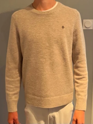 Beige stickad Morris tröja  - Klassisk beige stickad tröja med rund hals och ribbade muddar. Tröjan har en liten broderad detalj på bröstet och är tillverkad i ett mjukt material som passar perfekt till vardags. Långärmad och enkel design som är lätt att matcha.