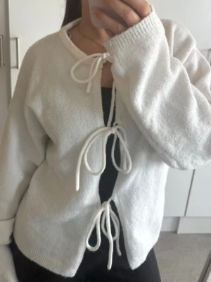 Vit kofta med knytband - Mysig vit kofta med tre knytband framtill. Koftan har rund halsringning, långa ärmar och är gjord i ett mjukt, stickat material. Perfekt för lager på lager och enkel att styla till olika outfits.