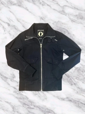 Cardigan från Elvine - Snygg mörkblå cardigan från Elvine med silverfärgad dragkedja, ribbade muddar vid ärmslut och midja samt två fickor med lock framtill. Jackan har en klassisk bomberkrage och detaljerade sömmar för en modern look.