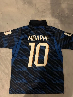 Frankrike fotbollströja Mbappé #10 - Säljer en blå och svart fotbollströja med Mbappé och nummer 10 på ryggen. Tröjan har korta ärmar, ståkrage och guldiga detaljer samt Frankrikes landslagsemblem på bröstet. Perfekt för dig som älskar fotboll och vill stötta Frankrike.