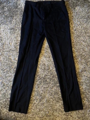 Svarta klassiska kostymbyxor - Snygga svarta kostymbyxor med rak passform och pressveck. Byxorna har bälteshällor, framfickor och två bakfickor med knapp. Perfekta för en stilren och clean look. Materialet ser ut att vara en mjuk och lätt tygblandning.
