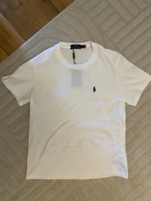 Vit t-shirt från Polo Ralph Lauren - Klassisk vit t-shirt från Polo Ralph Lauren med den ikoniska svarta logobrodyr på bröstet. T-shirten har rund hals och korta ärmar, tillverkad i mjuk bomull för en skön känsla. Perfekt basplagg till garderoben.