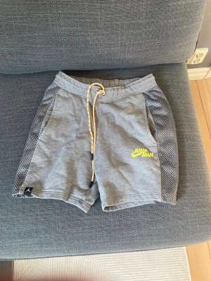 Grå shorts med snörning och meshdetaljer - Snygga grå shorts med meshdetaljer längs sidorna och gul 'Jumpman'-logga framtill. Shortsen har elastisk midja med gul/vit snörning och två fickor fram. Perfekta för träning eller chill dagar.