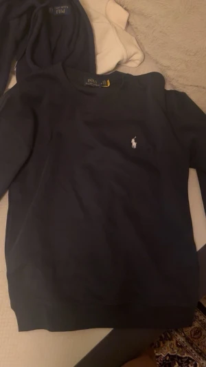Mörkblå stickad tröja Polo Ralph Lauren - Mörkblå stickad tröja från Polo Ralph Lauren med klassisk rund halsringning och den ikoniska vita logon broderad på bröstet. Tröjan har långa ärmar och ribbade muddar vid ärmslut och nederkant. Perfekt för en stilren och avslappnad look.