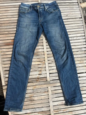 Blå slim fit jeans med slitningar från Jack & Jones i 31 32 - Snygga blå slim fit jeans med lätt slitna detaljer och klassisk femficksdesign. Jeansen har normalhög midja och smala ben. Tillverkade i denim med en tvättad look som ger en avslappnad känsla.