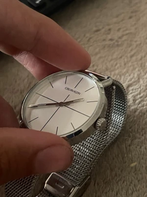 Silverfärgad klocka från Calvin Klein - Stilren klocka från Calvin Klein med rund, silverfärgad urtavla och mesh-armband i metall. Minimalistisk design med tunna svarta markeringar och silverfärgade visare. Perfekt accessoar för dig som gillar enkel och modern stil. Men sönder på sidan. Man kan inte snurra den!!