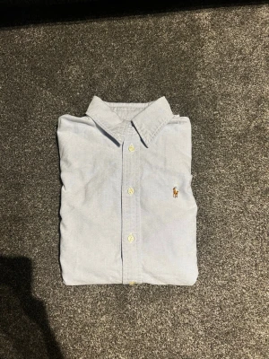 Ralph Lauren skjorta  - Klassisk ljusblå skjorta från Ralph Lauren med knappar framtill och broderad logga på bröstet. Skjortan har lång ärm och en tidlös design som passar till många olika stilar.