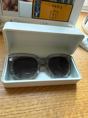 Gråa solglasögon från CHIMI - Snygga solglasögon från CHIMI med transparent grå båge och mörka glas. Modellen har en fyrkantig form och levereras i ett stilrent vitt fodral. Perfekta för att lyfta din look med en modern touch.