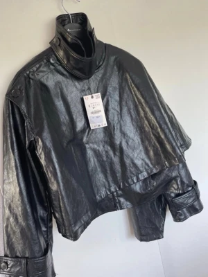 Svart jacka med hög krage från Zara - Snygg svart jacka  med hög krage och dekorativa knappar längs axlarna. Jackan har en boxig, croppad passform och breda ärmar. Perfekt för dig som vill ha en edgy och modern look. Stängs med knappar vid halsen och har coola detaljer.