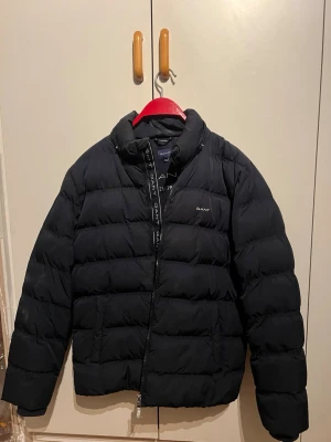 Svart pufferjacka från GANT - Säljer en svart pufferjacka från GANT med hög krage och dragkedja framtill. Jackan har diskret GANT-logga på bröstet och längs dragkedjan. Klassisk quiltad design och vadderad för extra värme. Perfekt för kalla vinterdagar. Storleken på jackan är 13-15 år/ 170cm