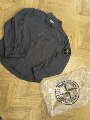 Svart overshirt från Stone Island - Snygg svart overshirt från Stone Island med dragkedja framtill, klassisk krage och bröstficka. Ikoniska Stone Island-loggan sitter på vänster ärm. Tillverkad i ett lätt, tekniskt material som ger en modern look.
