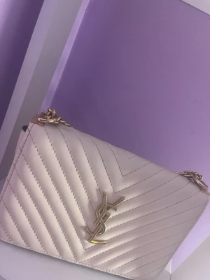Vit quiltad YSL handväska med kedja - Snygg vit quiltad handväska från YSL med guldfärgad kedjerem och klassisk YSL-logga framtill. Väskan har ett elegant mönster i v-format och detaljer i guld, perfekt för att lyfta vilken outfit som helst. Tillverkad i konstläder med både axelrem och kedja.