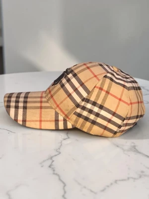 Beige rutig keps med Burberry-logga - Säljer en klassisk keps från Burberry i beige med det ikoniska rutmönstret i svart, vitt och rött. Kepsen har böjd skärm och en metallfärgad TB-logga framtill. Perfekt accessoar för en trendig och stilren look.