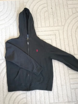 Svart hoodie från Polo Ralph Lauren - Svart hoodie från Polo Ralph Lauren med dragkedja och huva. Klassisk röd logga broderad på bröstet och kängurufickor framtill. Tillverkad i mjukt material som känns skönt mot huden. Perfekt för en chill och avslappnad stil. Stl 152