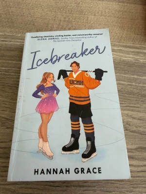 Icebreaker av Hannah Grace - En romantisk och humoristisk roman om konståkaren Anastasia och hockeykaptenen Nate. Boken har ett blått omslag med illustration av en tjej i rosa skridskoklänning och en kille i orange hockeytröja. Perfekt för dig som gillar sport, kärlek och ungdomsdrama.