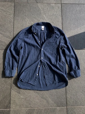 Mörkblå linneskjorta med knappar - Snygg mörkblå skjorta i linne med klassisk krage och vita knappar. Skjortan har en bröstficka och lång ärm, perfekt för en avslappnad men stilren look. Passar till både jeans och chinos.