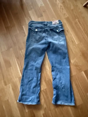 True Religion ljusblå bootcut jeans - Säljer ett par ljusblå jeans från True Religion med bootcut-modell. Jeansen har tydliga kontrastsömmar, stora bakfickor med lock och vita knappar. Klassisk patch bak i midjan och coola detaljer som sticker ut.
