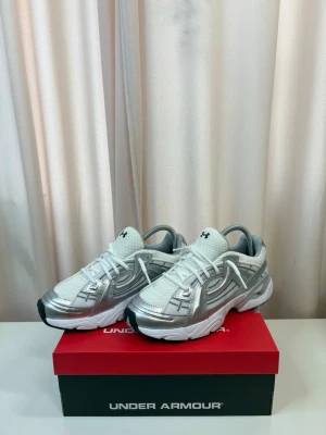 Under Armour Sola - Snygga sneakers från Under Armour i vitt och silver med futuristisk design. Ovandelen är i mesh och syntetmaterial med coola detaljer och logga på plösen. Tjock vit sula för extra komfort och sportig look. Perfekta för dig som gillar streetwear och sportig stil.